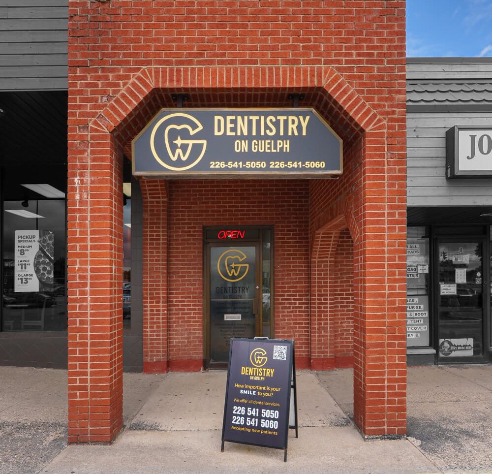 Front view of Dentistry on Guelph, a dental clinic with a brick exterior. The clinic's signboard is displayed above the entrance, showing the clinic's name and contact numbers. A sidewalk sign in front of the entrance provides additional information about the dental services offered and highlights the acceptance of new patients.