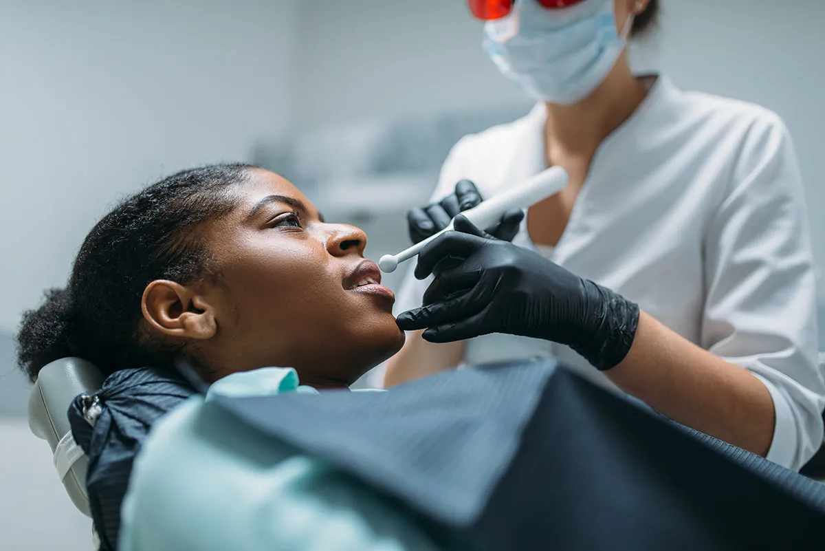 African American woman receiving dental treatment, lying in dental chair while dentist in white coat and mask performs examination with dental instrument