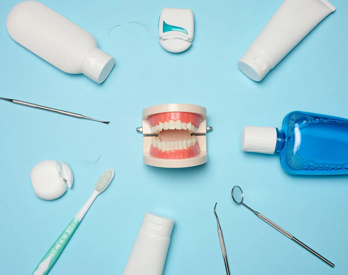 Dental hygiene products and tools arranged around jaw model on blue surface - toothbrush, floss, mouthwash, toothpaste tubes and dental examination instruments
