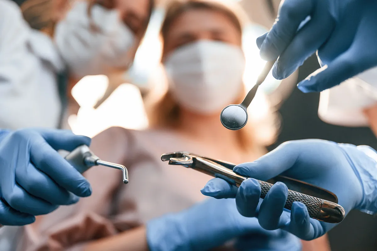 Close-up of dental procedure with multiple gloved hands holding dental instruments including mirror and drill, with masked dental team working on patient
