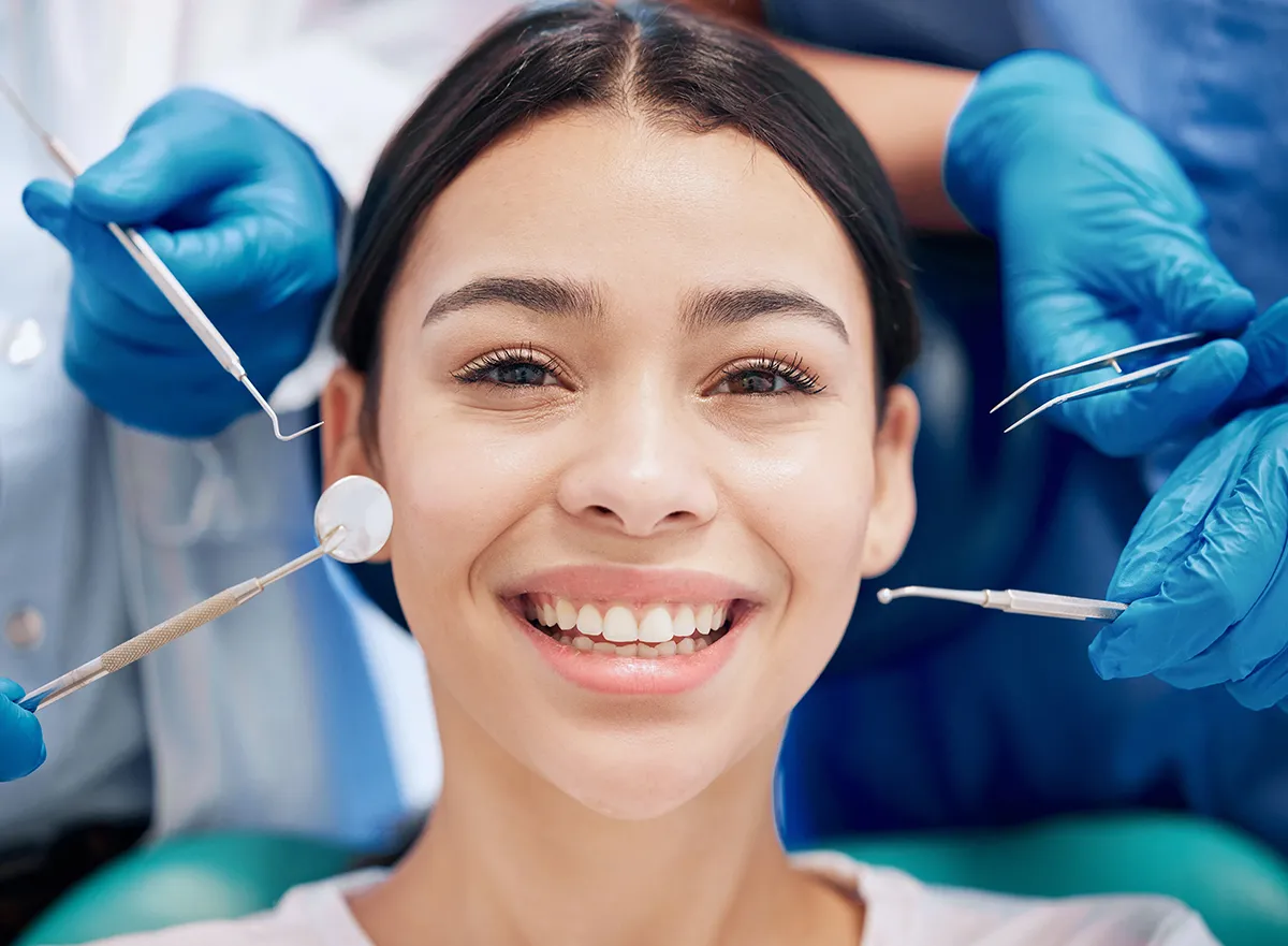 Smiling woman at dental office with dentist's gloved hands holding dental instruments including mirror and probe around her face
