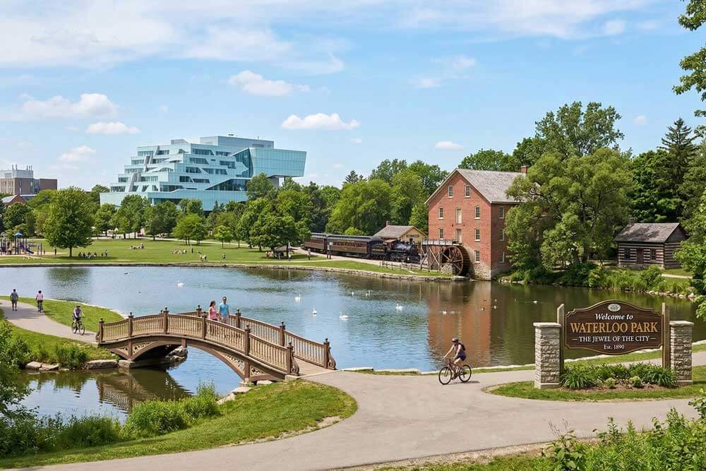 An idealized, AI-generated landscape of Waterloo Park in Ontario, Canada, featuring a scenic lake with white swans and a wooden arched bridge. The scene combines various local landmarks into one view, including the modern, glass architecture of the Perimeter Institute, a historic red brick grist mill with a water wheel, a vintage steam train, and a rustic log cabin. In the foreground, a stone sign reads "Welcome to Waterloo Park – The Jewel of the City – Est. 1890," while people walk and cycle along paved paths under a bright, sunny sky.