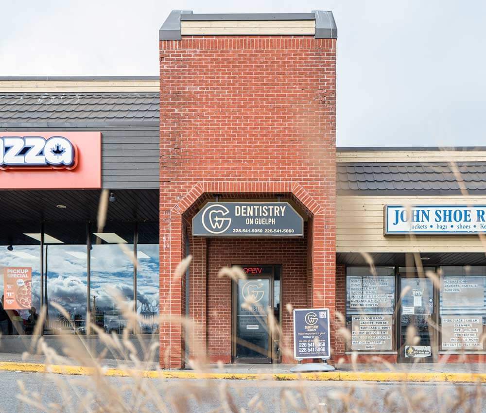 Exterior of "Dentistry on Guelph" dental clinic at 380 Eramosa Road. The storefront features a prominent red brick pillar archway with a black arched awning displaying the white "G" tooth logo and the business name.